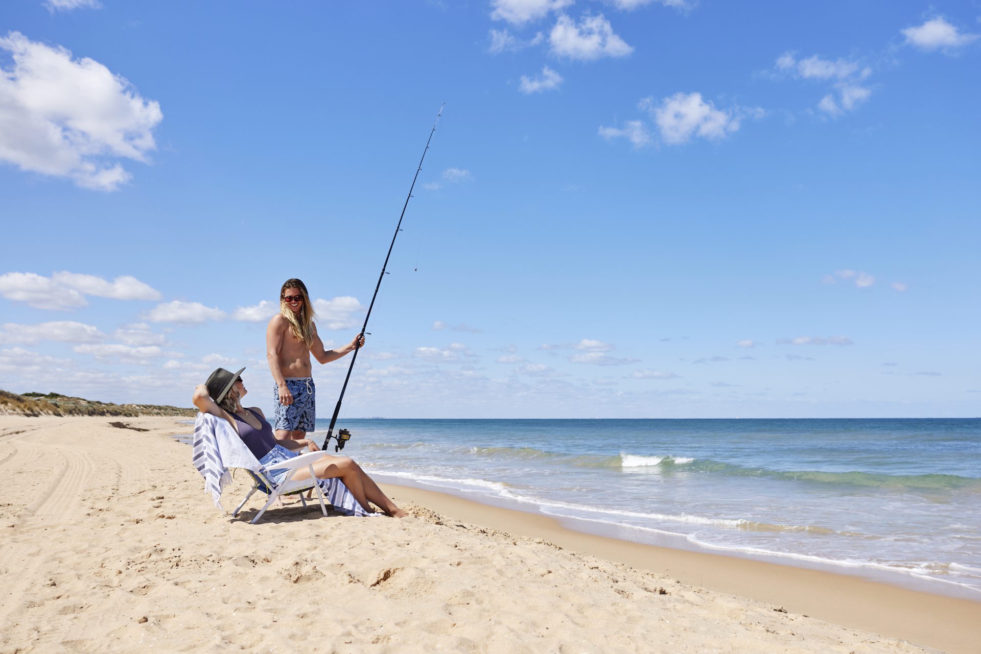 A man and a woman on the beach. The woman sits in a beach chair and the man stands next to her, holding a fishing rod.