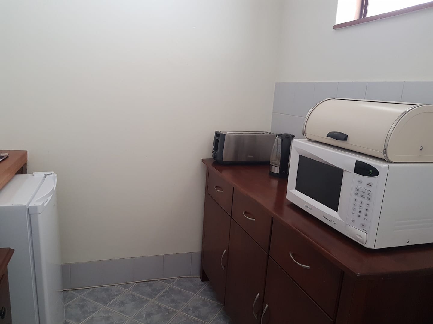 Small kitchenette with a toaster, kettle, microwave, bread bin and mini fridge.