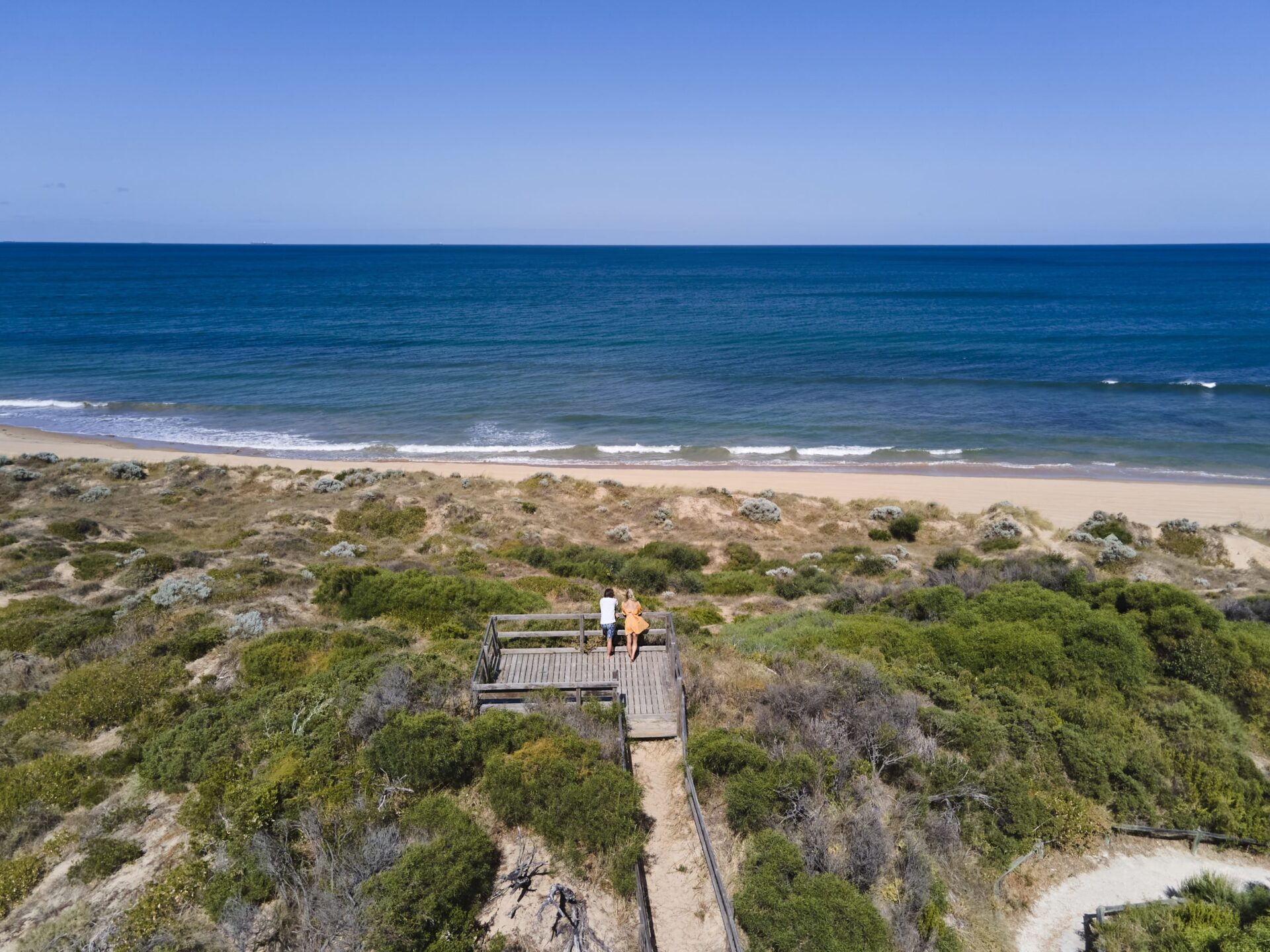 A drone image showing a man and woman standing on a wooden lookout, overlooking the beach and ocean.