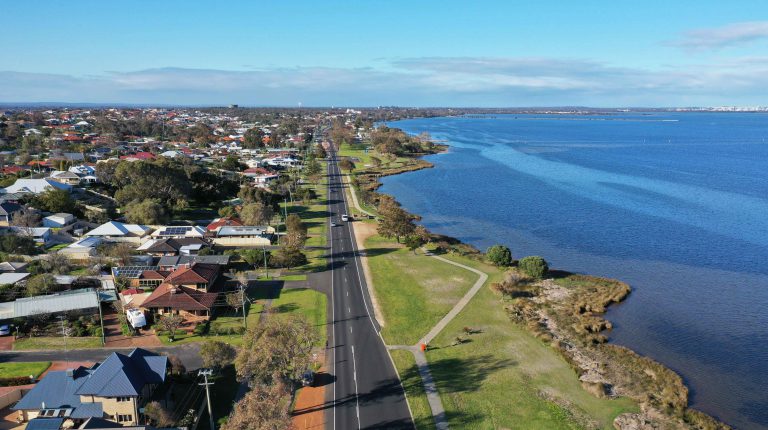 Drone image of a road. The road is in the middle of the image, with houses on the left and blue estuary water on the right. The top third of the image shows a blue sky with some clouds.