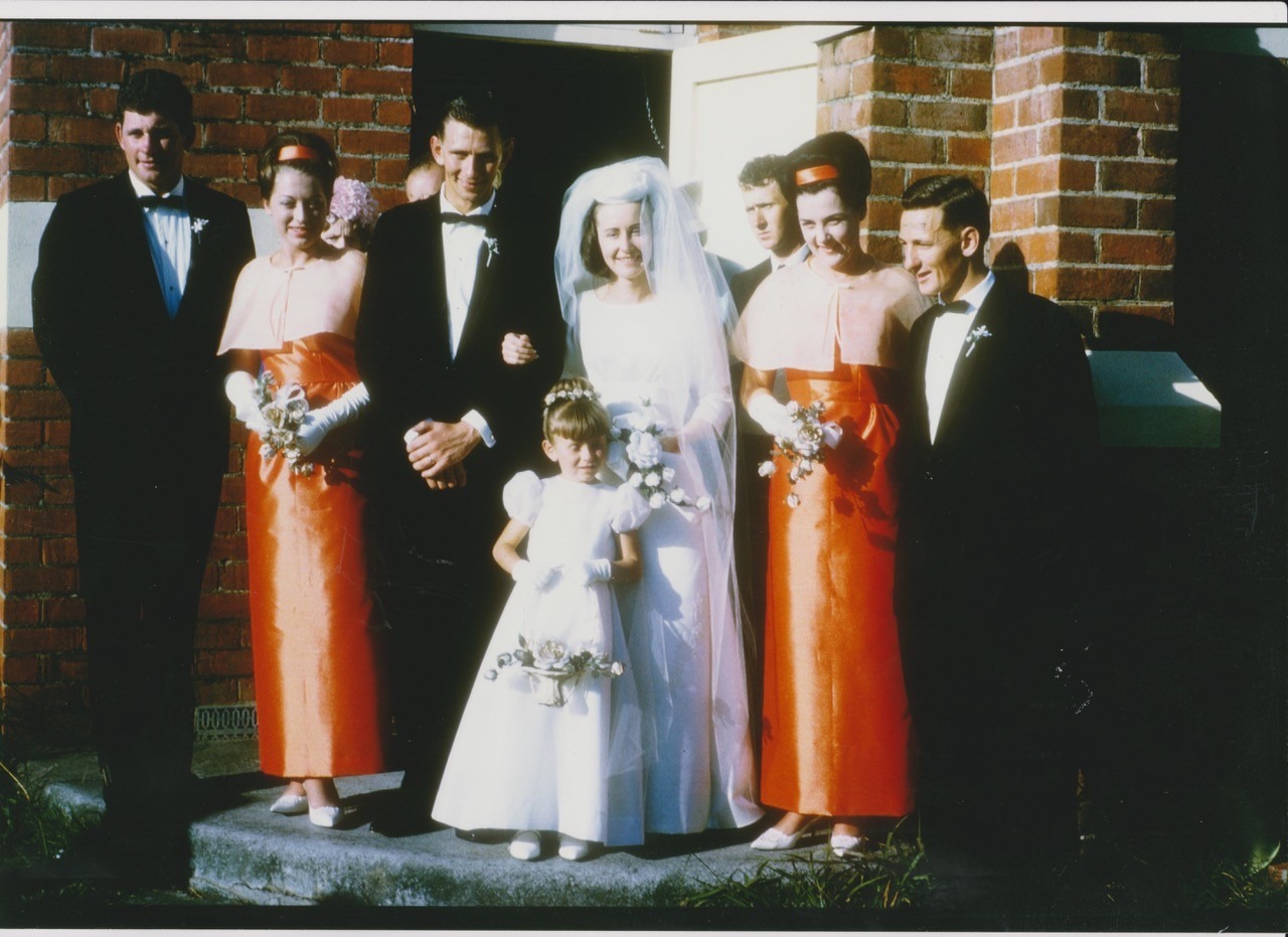 Old photo of wedding, men wearing black suits, bridesmaids in a burnt orange, bride in the middle in white with flower girl in front
